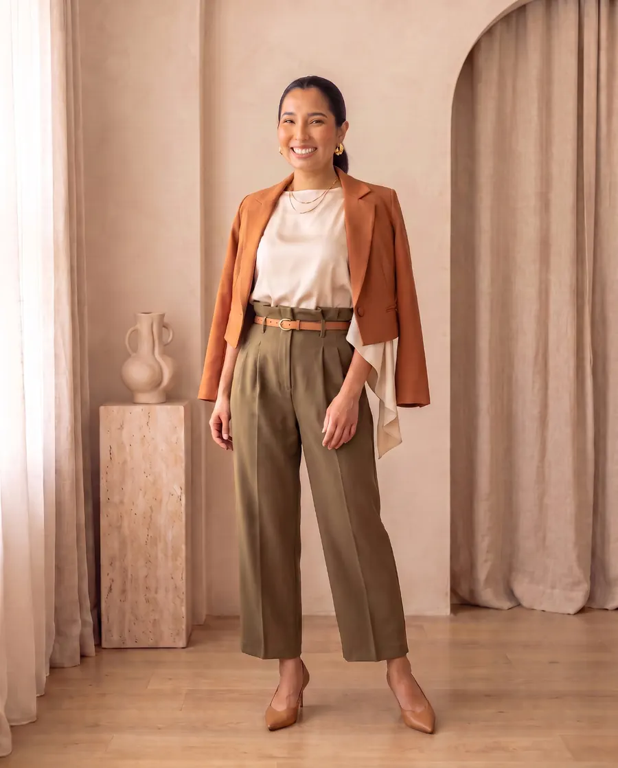 Helô Emy de corpo inteiro com blazer terracota, blusa creme e calça oliva, num cenário com cortinas de linho