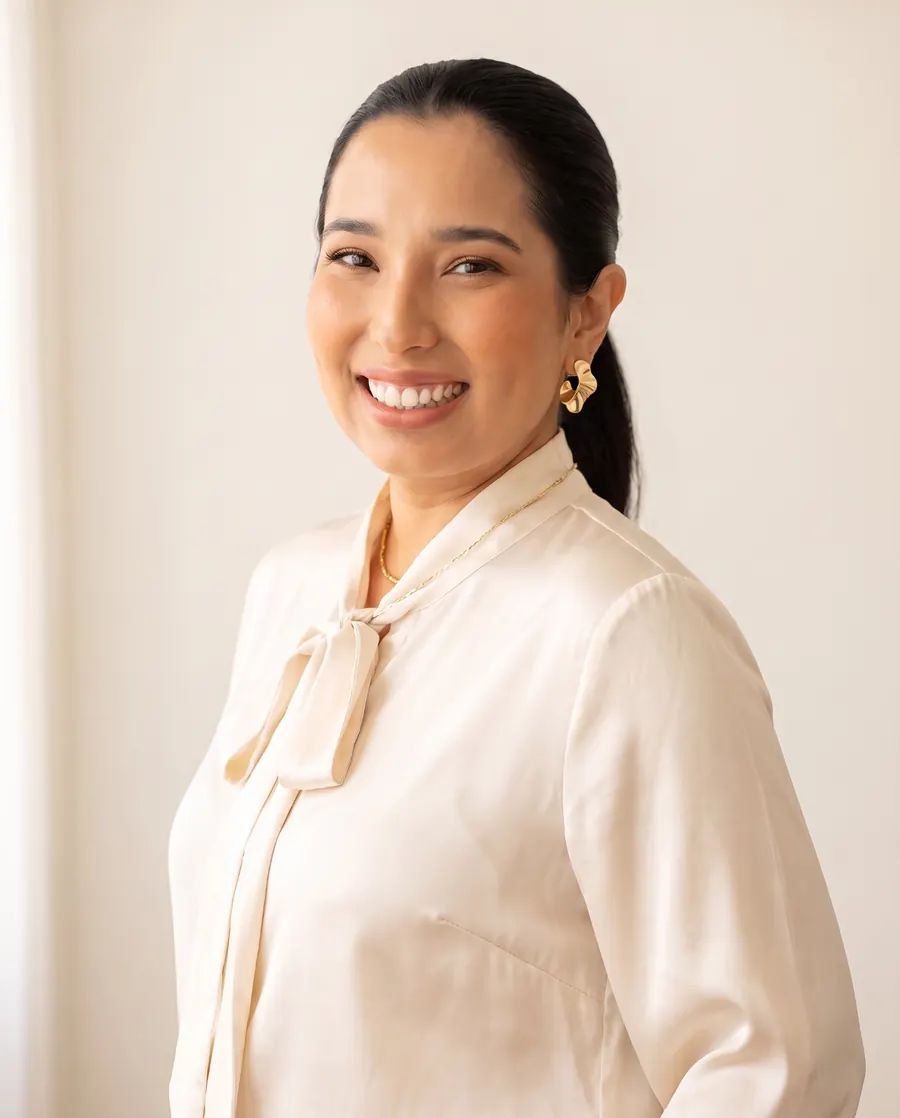 Retrato de Helô Emy sorrindo, com blusa de laço em cetim creme e brincos dourados